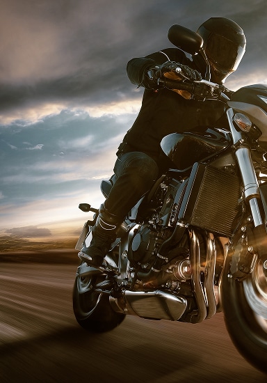 Motard casqué en tenue noire sur une moto de sport chromée en mouvement. Ciel nuageux et flou de vitesse dynamique sur la route.