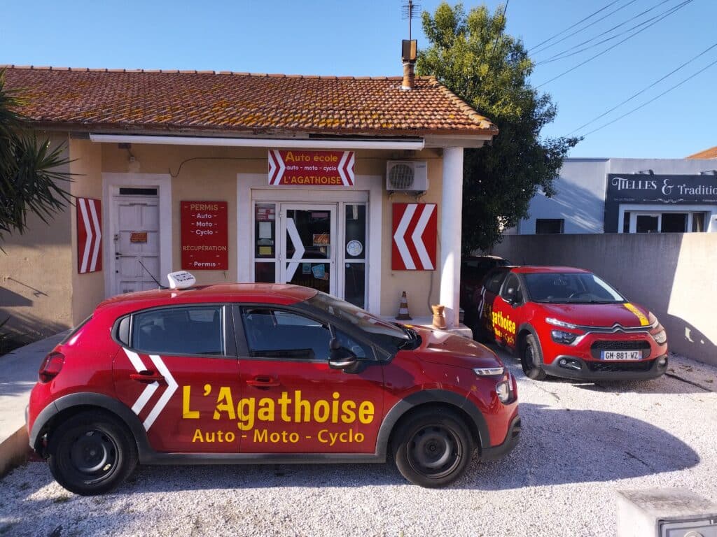 Bâtiment de l'auto-école L'Agathoise à Agde, avec deux voitures rouges et panneaux Auto, Moto, Cyclo, Permis.