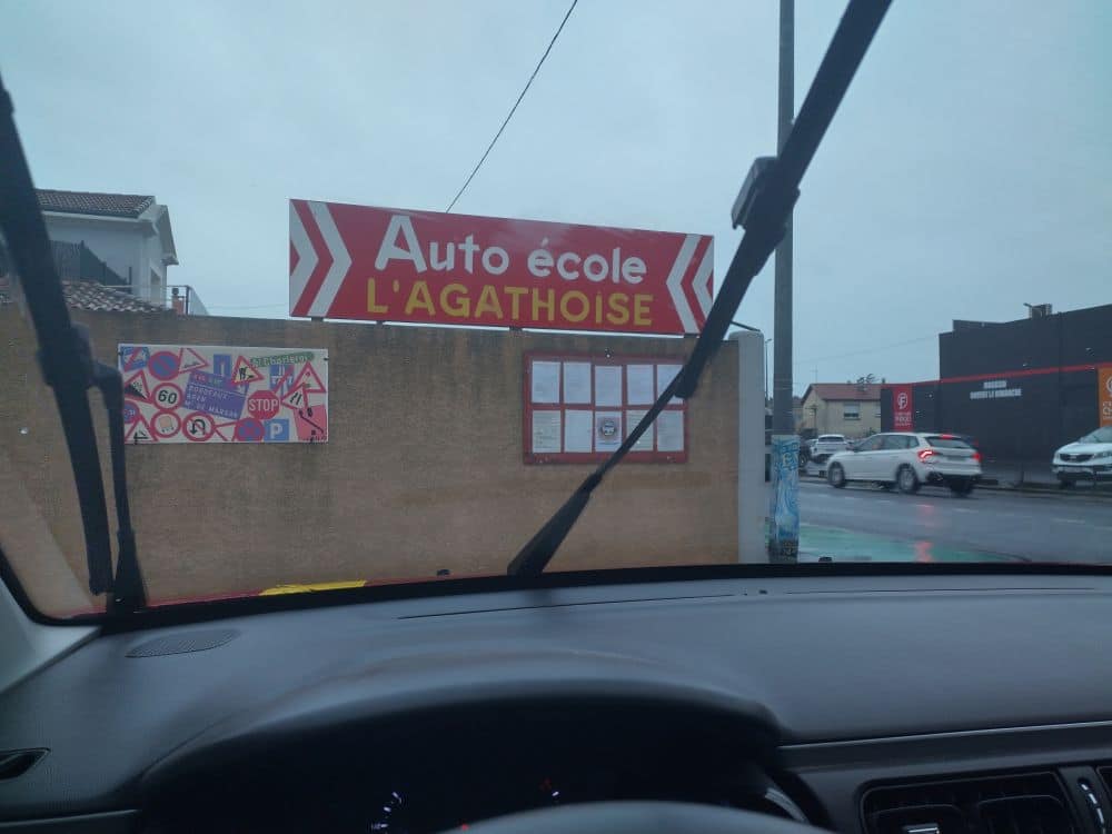 Panneau rouge de l'Auto école L'AGATHOISE vu à travers un pare-brise mouillé. Un mur expose des signaux routiers français variés (STOP, 60).