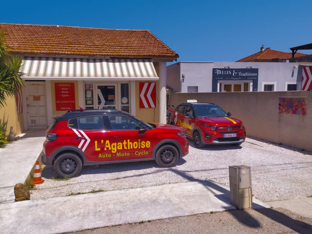 Auto-école L'Agathoise : Permis Auto, Moto et Cyclo Voitures rouges L'Agathoise Auto-Moto-Cyclo devant l'auto-école sous un ciel bleu.